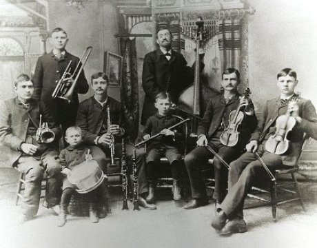 Das Schwarz/weiß Foto einer Band, der Bach Band in USA. 8 Personen, davon 7 Kinder unter 25. Alle haben Instrumente. Sechs sitzen, dahinter eine Orgel. In der Mitte hinten: Reinhold Bach mit Bratsche.