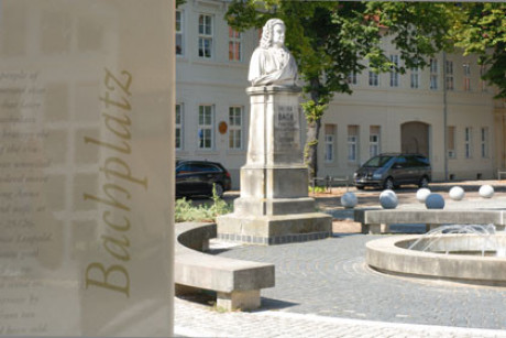 Im Foto sieht man das Bachdenkmal in Köthen ganz in weiß auf einem hohen Sockel, Bach bis zum Gürtel. Links sehr dominant im Vordergrund die Hinweistafel zum Bachplatz. Dahinter ein sehr großes Haus.