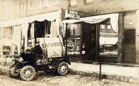 Es ist ein historisches schwarzweißes, leicht vergilbtes Foto mit einem sehr historischen Lkw vor einem Ladengeschäft in den USA. Auf dem Geschäft und auf dem Lkw liest man „Bach“.