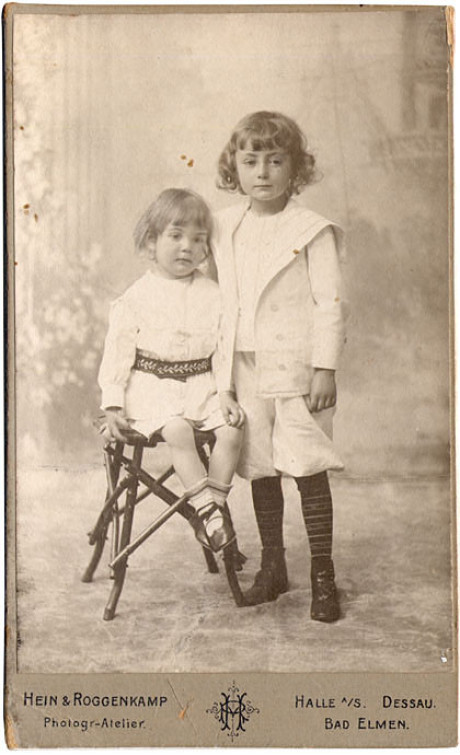 Auf einem historischen hochformatigen Bild schauen zwei Kinder zum Betrachter. Das Kind rechts ist einen Kopf größer als das Kind links, das auf einem Hocker sitzt. Links ist es ein Mädchen, rechts ist es ein Junge. Das Foto ist leicht verbleicht. Der Hin