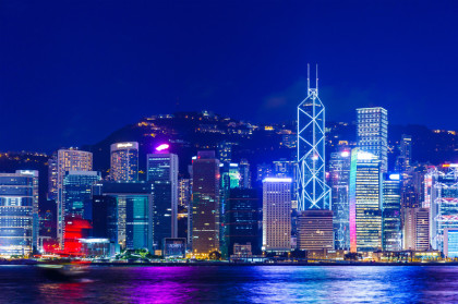 Im Bild sieht man die Wolkentratzer-Skyline von Hongkong. Auch in Honkong gibt es einen Bachchor. Hinter den Hochhäusern sieht man einen Berg, vorne ist Gewässer. Blau überwiegt in diesem Nachtmotiv, der Himmel ist dunkles royalblau.