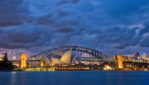 Das Bild zeigt die Skyline von Sydney mit der Oper bei Nacht. Der Himmel ist stark bewölkt. Auch Sidney hat einen Bachchor. Hinter der Oper sieht man die berühmte Brücke. Die Oper ist beleuchtet. Das komplette Foto hat einen Blau-Touch.