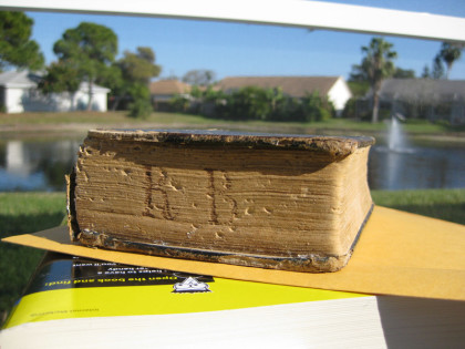 Auf dem Foto sieht man das uralte Gesangbuch schräg fotografiert auf einem Dummy-Buch zum späteren Nachbearbeiten, im Hintergrund ein See. Man sieht die Buchstaben R und B auf den Seiten eingeritzt.