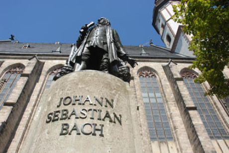 Im Bild ist das Denkmal von Bach vor der Thomaskirche imposant extrem von nah und unten fotografiert: der Schriftzug extrem groß, der Kopf weit entfernt. Im Hintergrund die Kirche, blauer Himmel.