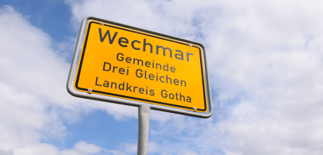 Man sieht im Bild zum Kapitel Ursprung drei aktuelle Wohnhäuser im Hintergrund. Der Fokus liegt auf dem gelben Ortseingangsschild und dem Verkehrszeichen Vorfahrt. Fotografiert ist aus einem Kornfeld.