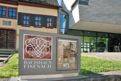 Links vorne sind die Bach-Insignien (Bach-Logo), weiß auf rostrotem Grund, sehr groß in einem Glaskasten vor dem Bachhaus und dem Bach-Museum in Eisenach. Der Kasten ist etwa einen Meter hoch und zwei Meter breit und man sieht dort im rechten Teil weitere