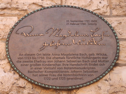 Die Plakette auf dem Thomashof für Anna Magdalena Bach. Sie ist bronzefarben und im Hintergrund sieht man das Gemäuer der Kirche. Darauf ist ihr Name als Anna Magdalena Bachin, geborene Wülckin. Darunter sind einige Hinweise.