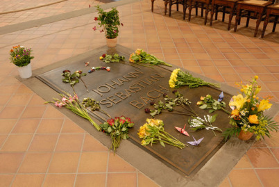 Die Grabplatte im vorderen Teil der Thomaskirche Man liest in großen Lettern und in drei Zeilen: Johann Sebastian Bach. Viele Blumensträuße liegen auf dem Grab und dort steht auch noch ein Blumengesteck. Im Hintergrund sieht man Stühle.