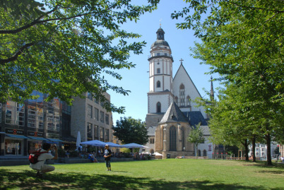 Die Thomaskirche in der Bildmitte, zwar von der Rückseite, aber es ist die bekanntere als die Vorderseite. Vorne ist eine Rasenfläche, auf der ein Vater seinen Sohn fotografiert, rechts und links sind Bäume. Links dahinter auch Häuser.