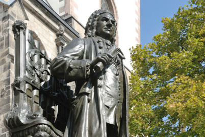 Das Alte Bach-Denkmal. Rechts ist Johann Sebastian Bach und hält gerolltes Notenpapier in der Hand, links ist die Orgel hinter ihm zu sehen. Links ist ein Teil der Thomaskirche im Sonnenschein sichtbar, rechts ein Baum im Frühlingslaub vor blauem Himmel.