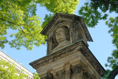 Das Alte Bach-Denkmal, von unten nach oben fotografiert und zwar gegen einen blauen Himmel ohne Wolken und hellgrünes Frühlingsblattwerk. Fotografiert ist der obere Teil des rund 3 Meter hohen Monuments mit Bachs Büste an einer der 4 Seiten.
