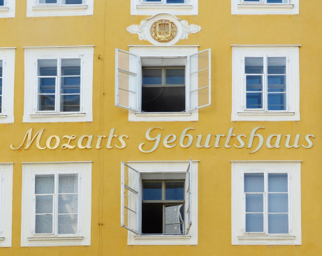Man sieht ein gelben Haus, den Auschnitt der Frontseite mit sechs weißen Sprossenfenstern. Die mittleren Fenster sind offen, das Haus ist gelb und dort steht Mozarts Geburtshaus.
