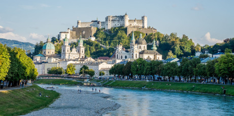 Auf einem Berg vor einem Fluß in der unteren Bildhälfte liegt Salzburgs Schloss und darunter die Satdt. Links sind Bäume im Frühling, der Himmel ist fast wolkenlos.
