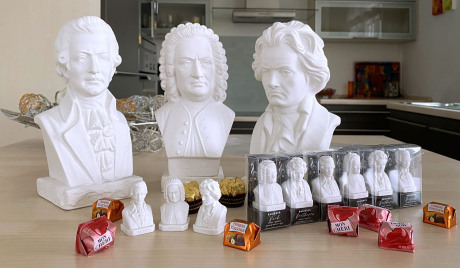 Auf einer modernen Kücheninsel stehen drei große und neun winzige Bach-, Mozart- und Beethoven-Büsten sowie einige Süßigkeiten als Größenvergleich. Im Hintergrund sieht man die Küche.