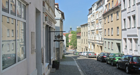 In der Stadt Zeitz sieht man einen Straßenzug, der leicht zum Horizont hin abfällt. Im Hintergrund ist der Turm des Stadtschlosses zu sehen. Vorne links, kaum wahrnehmbar, ist die Gedenktafel für Anna Magdalena Bach an einer Hauswand.
