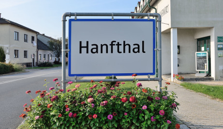 Es ist ein Ortseingangsschild nach Hanfthal. Links führt eine Straße in den Hintergund. Links und rechts sind Häuser. Vor dem Schild sind üppig schöne Blumen. Der Himmel ist blau.