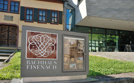 Das Bachsiegel in einem verglasten Schaukasten im Freien vor dem Bachhaus und Bach-Museum in Eisenach. In der linken Hälfte ist das Bach-Siegel ohne Krone und ohne Lorbeerzweige darunter. Es ist weiß auf braunem Grund. Darunter steht „Bachhaus Eisenach“.