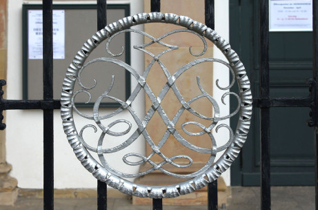 An einem vergitterten Tor vor einer Türe ist das Bachsiegel in Silber zu sehen. Es ist mit einem runden Gefelcht eingerahmt. Das Tor besteht aus schwarzen Gitterstäben.