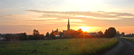 Über eine Wiese links und einen Schotterweg rechts sieht man in mittlerer Entfernung den Kirchturm von Wechmar, einige Häuser rechts und links und die aufgehende Sonne.
