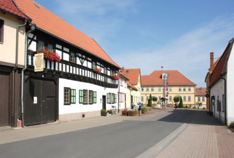 In die Tiefe des Bildes führt eine Straße auf den Gasthof „Goldener Löwe“ zu. Links steht das Bachstammhaus, ein Fachwerkhaus. Die Sonne scheint, der Himmel ist blau.