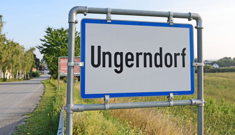Groß im Bild und dominant ist das Ortseingangsschild von Ungerndorf in Österreich in Blau und Weiß. In die Bildtiefe verläuft eine Straße. Man sieht Bäume links und einen Acker rechts.