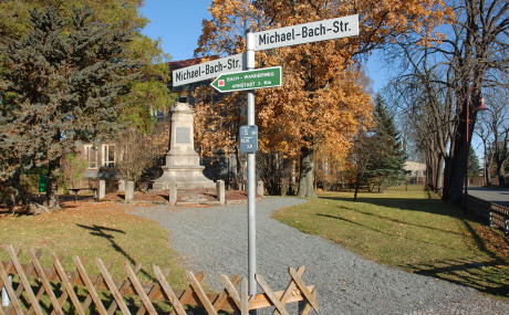 Neben einem Gartenzaun führt ein Weg zu einem Denkmal in Form eines Obelisken. Rechts und links sind Bäume. Mittig ist ein Schild, das den Namen der Straße verrät.