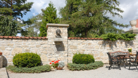 Vor einer runden Bepflanzung links und einer Gartensitzgruppe rechts ist eine Mauer, die das Bachdenkmal in Dornheim integriert. Darunter ist eine Tafel mit Hinweis auf die Hochzeit von Maria Barbara Bach und Johann Sebastian Bach.