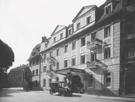Es ist ein historisches Schwarz/Weiß-Foto. Man sieht ein  vierstöckiges Haus, offensichtlich ein Hotel mit einem historischen schwarzen Kfz davor. Im Hintergrund sind weitere Häuser, vorne links ist die Straße.