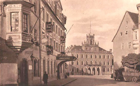 Links sieht man ein Hotel mit einem dominanten Erker, auf das man im steilen Wikel schaut. In der Bildmitte im Hintergrund ist das Rathaus von Weimar. Rechts sieht man einen Baum und einen Teil eines weiteren Hauses.